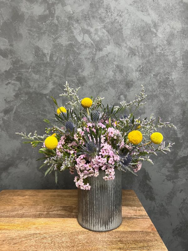 Delicate flower arrangement of yellows and purples in tall metal vase.