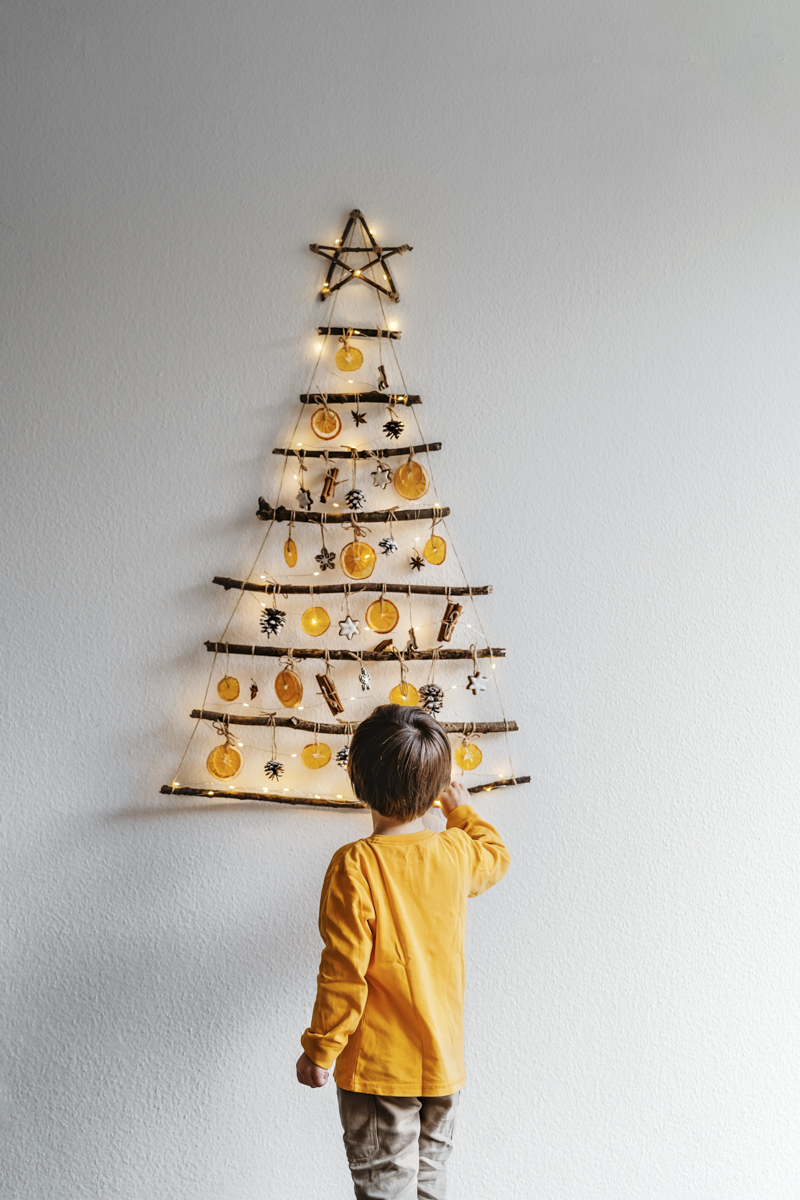 Little child decorating handmade craft Christmas tree made from sticks and natural materials hanging on wall. 
