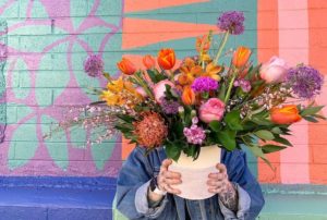 Person holding colorful flower arrangement in a white vase in front of painted wall..