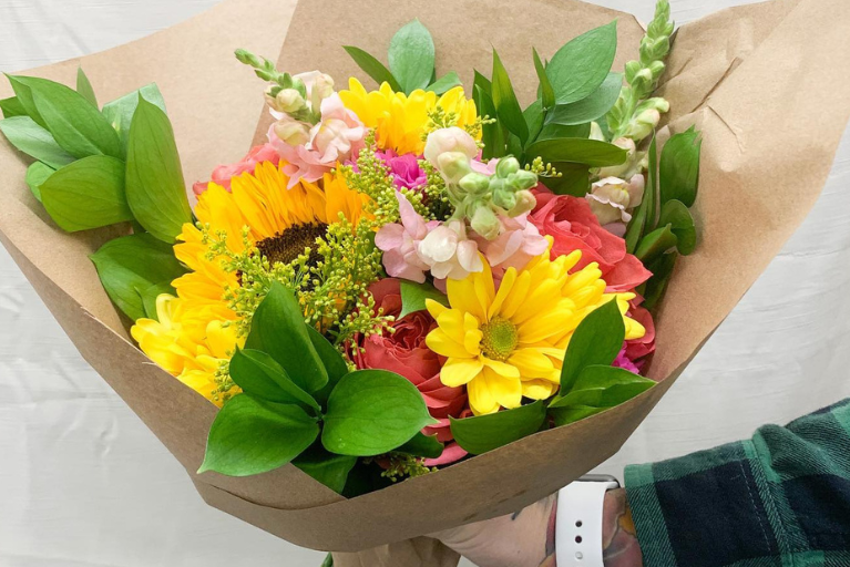 Arm holding a paper wrapped flower arrangement of multi color flowers for delivery.