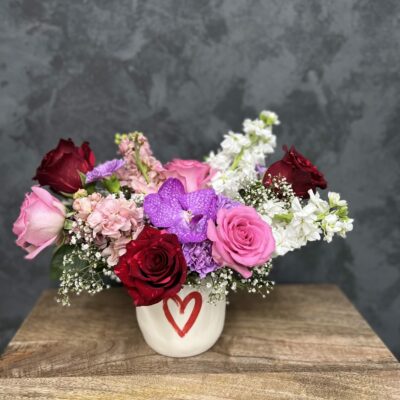 Valentine's flower arrangement of pinks and purples in a white vase with a red heart graphic.