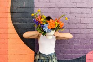 Person holding a vase of flowers in front of their head with a colorful brick wall in the background.