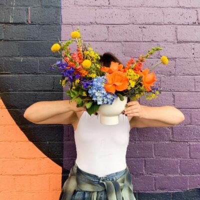 Person holding a vase of flowers in front of their head with a colorful brick wall in the background.