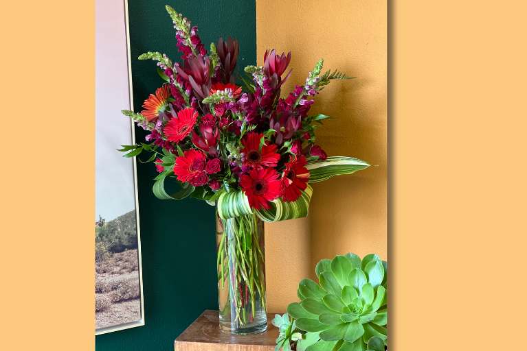 Red daisies and other flowers and green in a clear glass vase on a table.