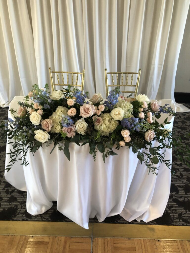 Grouping of pastel colored flowers and greens on a table covered with a white tablecloth.