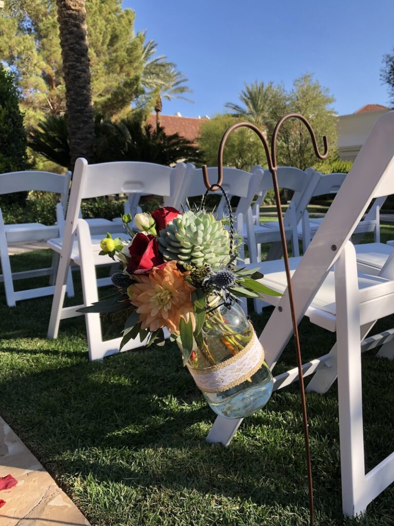 Glass vase of colorful flowers hanging from a hook at an event set up with white chairs.