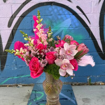 colorful flower arrangement of pink flowers and feathers in a shiny vase sitting on the ground in front of an art mural